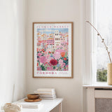 a framed poster of a colorful floral scene hanging on a wall, with a white table and a vase of flowers nearby.