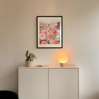 a white cabinet with a potted plant and a lamp on top, set against a wall with a framed poster of a cityscape hanging above it.