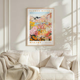 a framed poster of a colorful floral scene hanging on a wall above a white couch with a blanket draped over it.