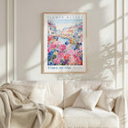 a framed poster of a colorful floral scene hanging on a wall above a white couch with a blanket draped over it.