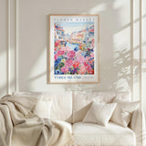 a framed poster of a colorful floral scene hanging on a wall above a white couch with a blanket draped over it.