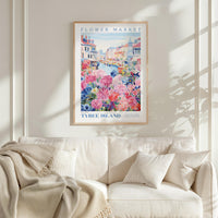 a framed poster of a colorful floral scene hanging on a wall above a white couch with a blanket draped over it.