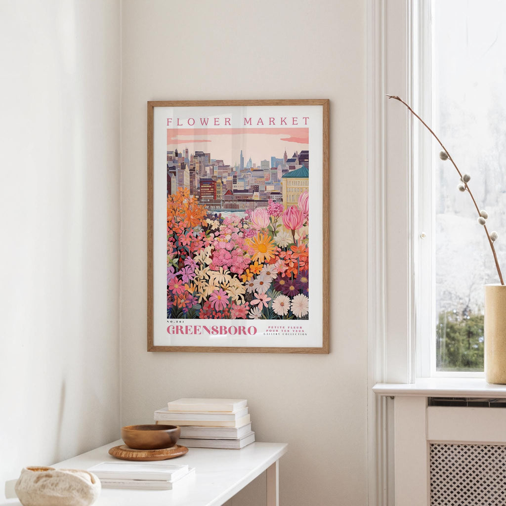 a framed poster of a colorful cityscape with flowers hanging on a wall in a room with a white table and a window.