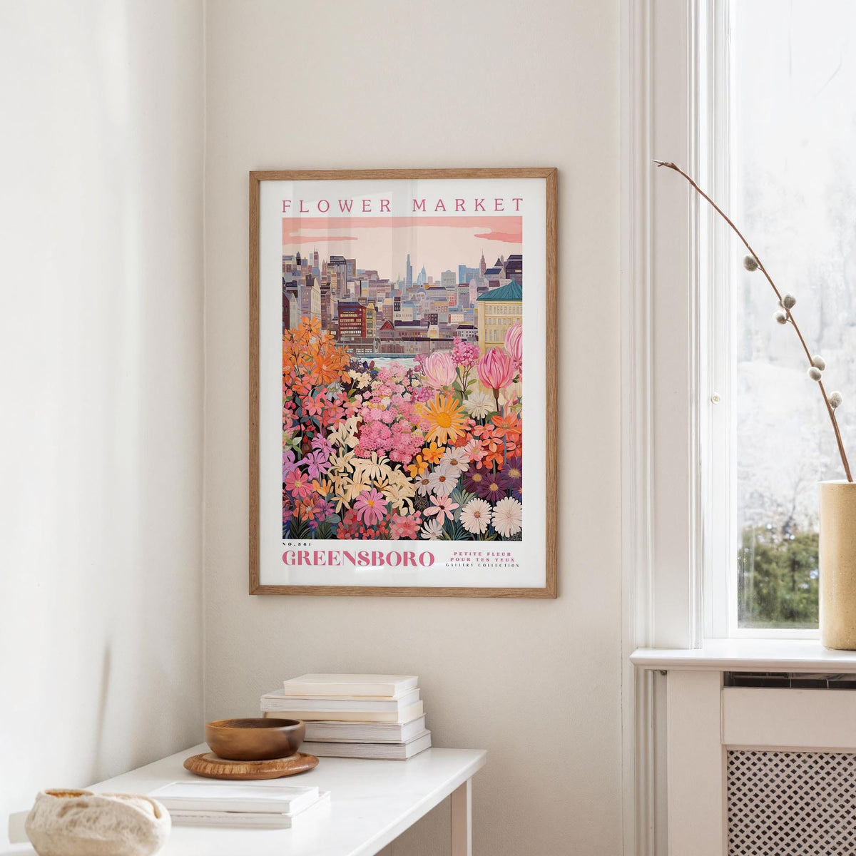 a framed poster of a colorful cityscape with flowers hanging on a wall in a room with a white table and a window.