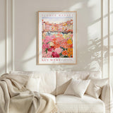 a framed poster of a colorful floral scene hanging on a wall above a white couch with a blanket draped over it.