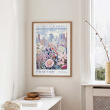 a framed poster of a cityscape with a flower market in Chicago, hanging on a wall in a room with a window and a small table with books and a vase.
