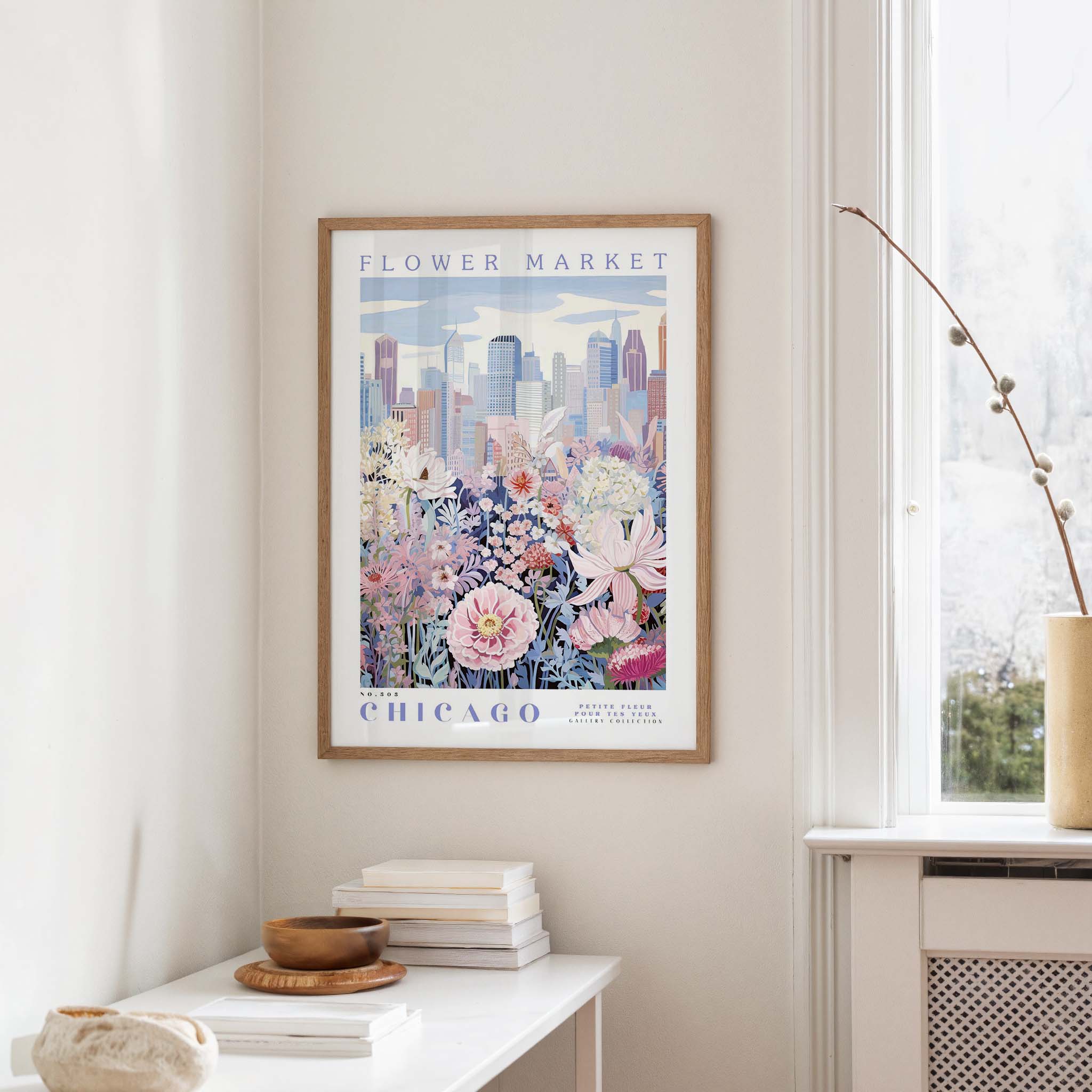 a framed poster of a cityscape with a flower market in Chicago, hanging on a wall in a room with a window and a small table with books and a vase.