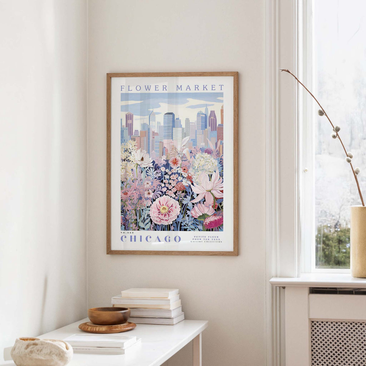 a framed poster of a cityscape with a flower market in Chicago, hanging on a wall in a room with a window and a small table with books and a vase.