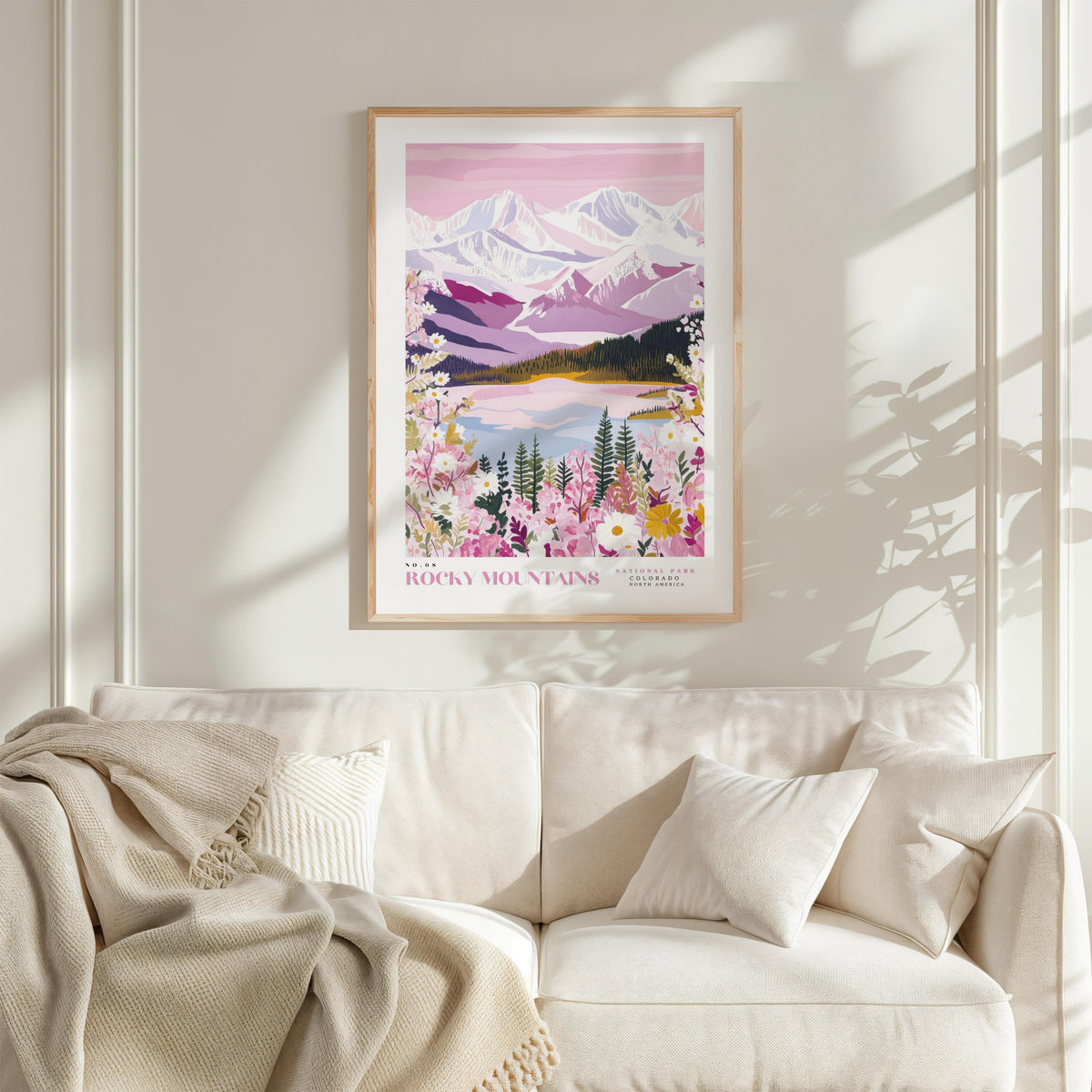 a framed poster of a mountain landscape with a lake and flowers hanging on a wall above a white couch with a blanket draped over it.