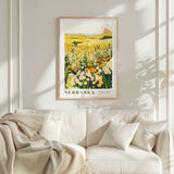 a framed poster of a field of flowers hanging on a wall above a white couch with pillows.