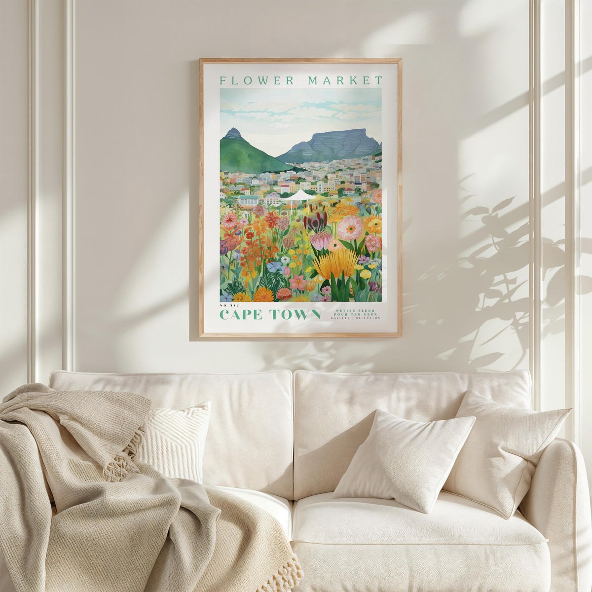 a framed poster of a colorful flower market in Cape Town, South Africa, hanging on a wall above a white couch.
