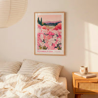 a bedroom scene with a framed poster of a field of pink flowers hanging on the wall above a bed with striped pillows.