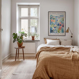 a cozy bedroom with a large bed, a wooden nightstand, and a potted plant. The room has a window with a view of a garden, and a colorful poster on the wall.
