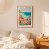 a framed poster of a coastal landscape with a flower market, hanging on a wall above a bed with striped pillows.