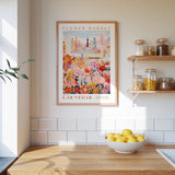 a framed poster of a colorful cityscape with a flower market in the background, hanging on a wall above a wooden counter with a bowl of lemons.