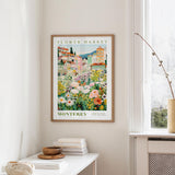 a framed poster of a colorful flower market in Montreya, Spain, hanging on a wall in a room with a window and a table with books and a vase.
