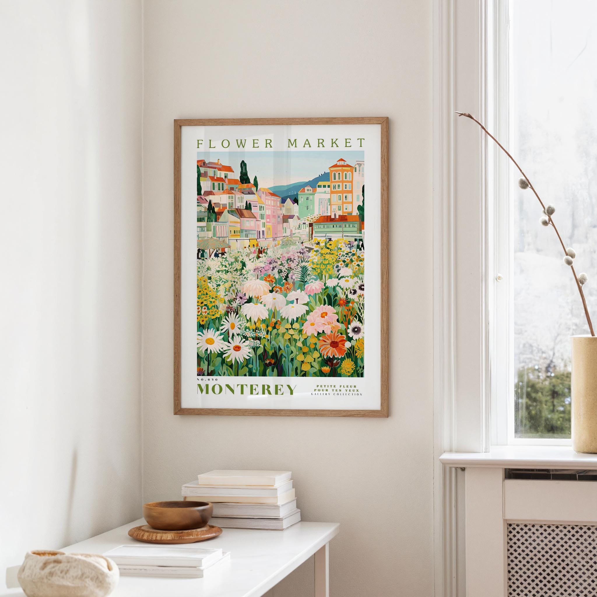 a framed poster of a colorful flower market in Montreya, Spain, hanging on a wall in a room with a window and a table with books and a vase.