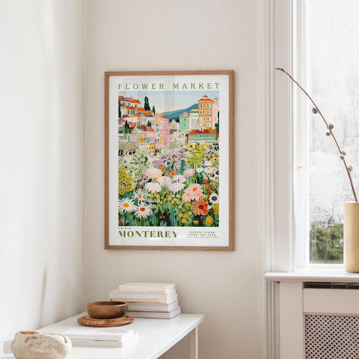 a framed poster of a colorful flower market in Montreya, Spain, hanging on a wall in a room with a window and a table with books and a vase.