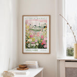 a framed poster of a flower market in Adelaide, Australia, hanging on a wall in a room with a window and a small table with books and a vase.