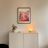 a white cabinet with a potted plant and a lamp on top, set against a white wall with a framed poster of a flower market above it.