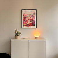 a white cabinet with a potted plant and a lamp on top, set against a white wall with a framed poster of a flower market above it.