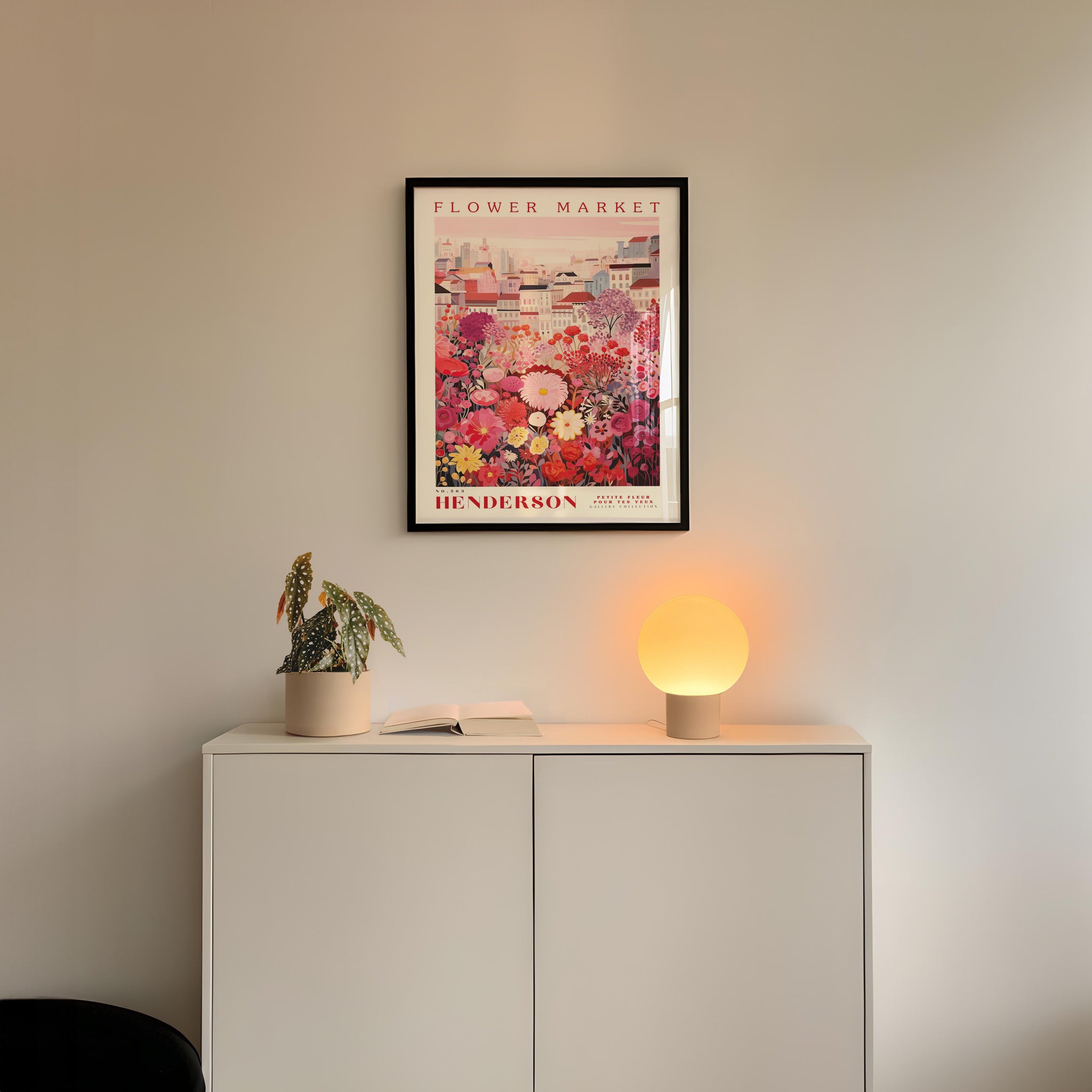 a white cabinet with a potted plant and a lamp on top, set against a white wall with a framed poster of a flower market above it.