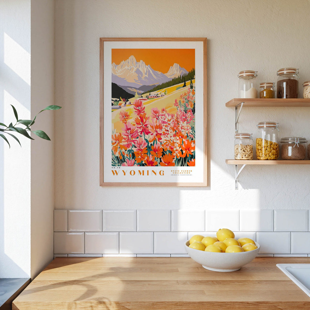 a framed poster of a scenic mountain landscape with a field of flowers, hanging on a wall above a kitchen counter with a bowl of lemons and other kitchen items.