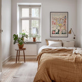 a cozy bedroom with a large bed, a wooden nightstand, and a window with a plant on the sill. The room has a warm, inviting atmosphere with a neutral color palette and natural elements like the potted plant and the rug.