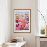 a framed poster of a colorful floral scene hanging on a wall, with a white table and a vase of flowers in the foreground.