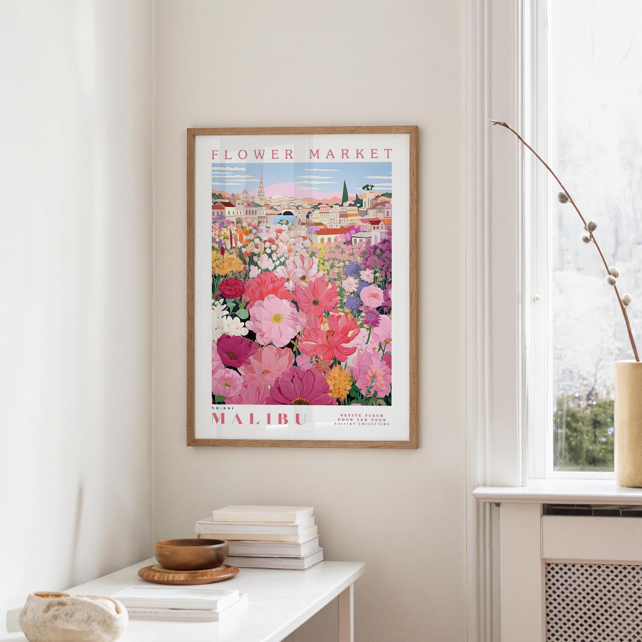 a framed poster of a colorful floral scene hanging on a wall, with a white table and a vase of flowers in the foreground.
