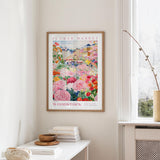 a framed poster of a colorful floral scene hanging on a wall, with a white table and a vase of flowers nearby.