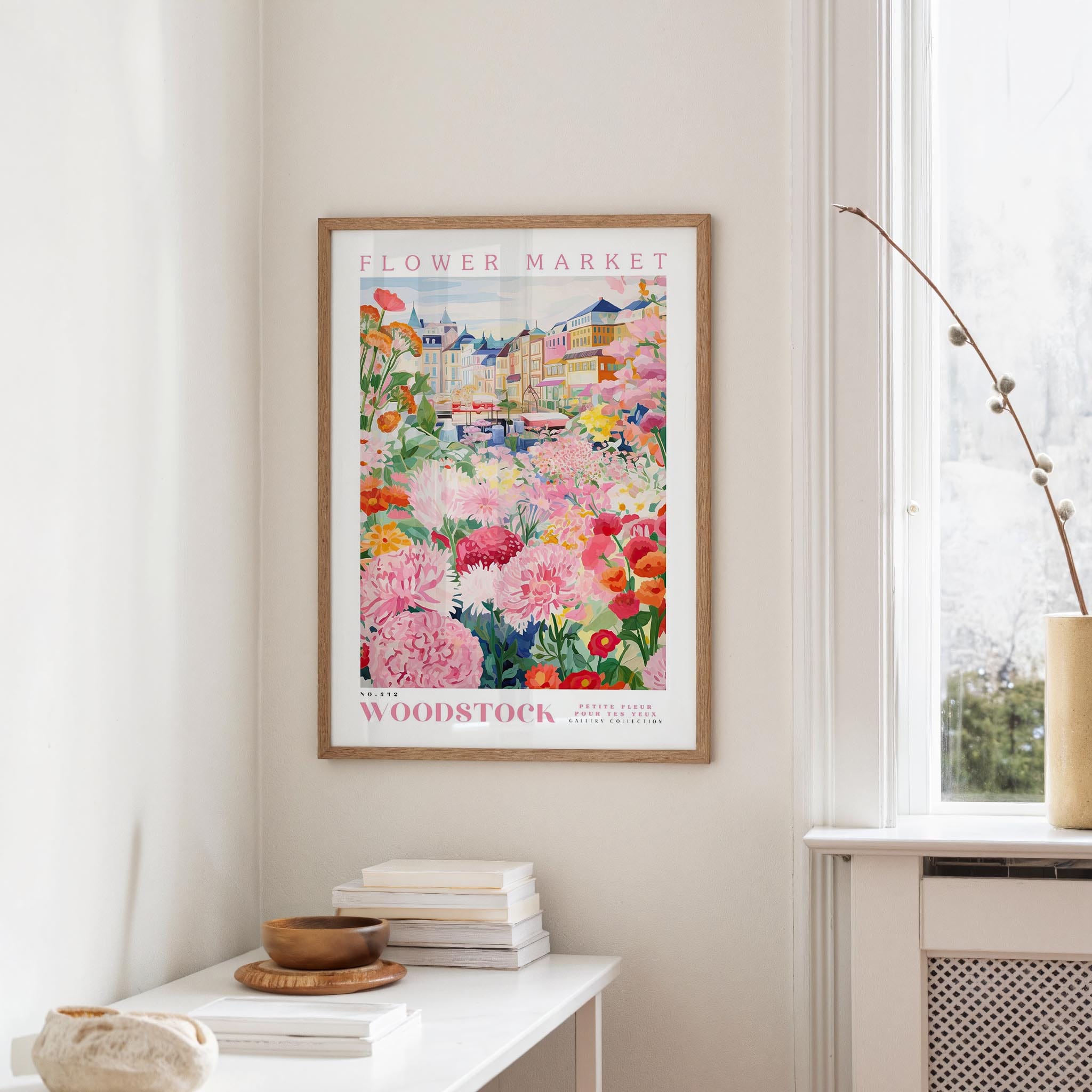 a framed poster of a colorful floral scene hanging on a wall, with a white table and a vase of flowers nearby.