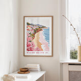 a framed poster of a beach scene with a coastal town in the background, hanging on a wall in a room with a window and a small table with books and a bowl on it.