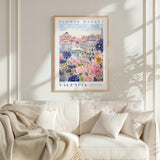 a framed poster of a colorful floral scene hanging on a wall above a white couch with a blanket draped over it.