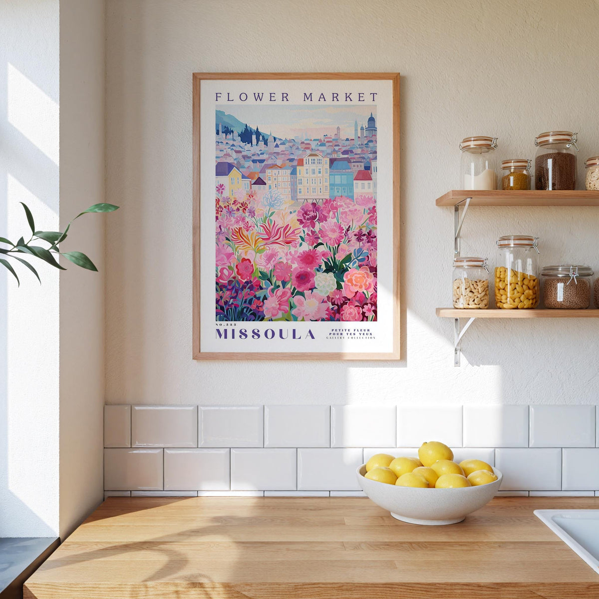 a kitchen scene with a framed poster of a flower market in Missoula, Montana, hanging on the wall above a wooden counter. The poster displays a colorful illustration of flowers and buildings, and the text "FLOWER MARKET MISSOULA" is visible. On the counter, there is a bowl of lemons, and the wall has white tiles.