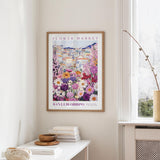 a framed poster of a colorful floral scene hanging on a wall, with a white table and a vase of flowers nearby.