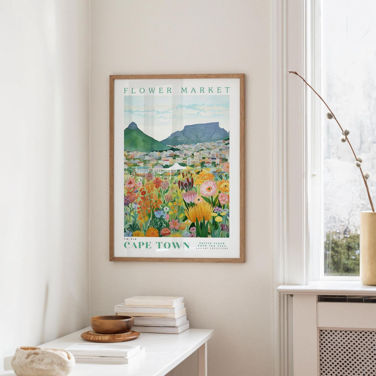 a framed poster of a colorful flower market in Cape Town, with mountains in the background. The poster is hanging on a wall, and there is a table with books and a vase nearby.