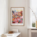 a framed poster of a colorful cityscape with flowers hanging on a wall, next to a window and a small table with books and a bowl.