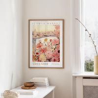 a framed poster of a cityscape with a floral design, hanging on a wall in a room with a white table and a window.