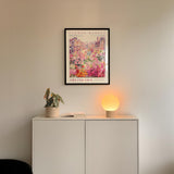 a white cabinet with a potted plant and a lamp on top, set against a wall with a framed poster of a flower market above it.