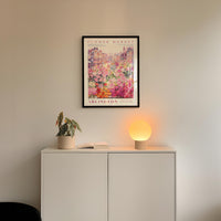a white cabinet with a potted plant and a lamp on top, set against a wall with a framed poster of a flower market above it.