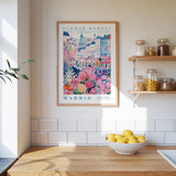 a framed poster of a colorful illustration of a flower market in Madrid, Spain, hanging on a wall above a wooden counter with a bowl of lemons.
