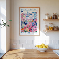 a framed poster of a colorful illustration of a flower market in Madrid, Spain, hanging on a wall above a wooden counter with a bowl of lemons.