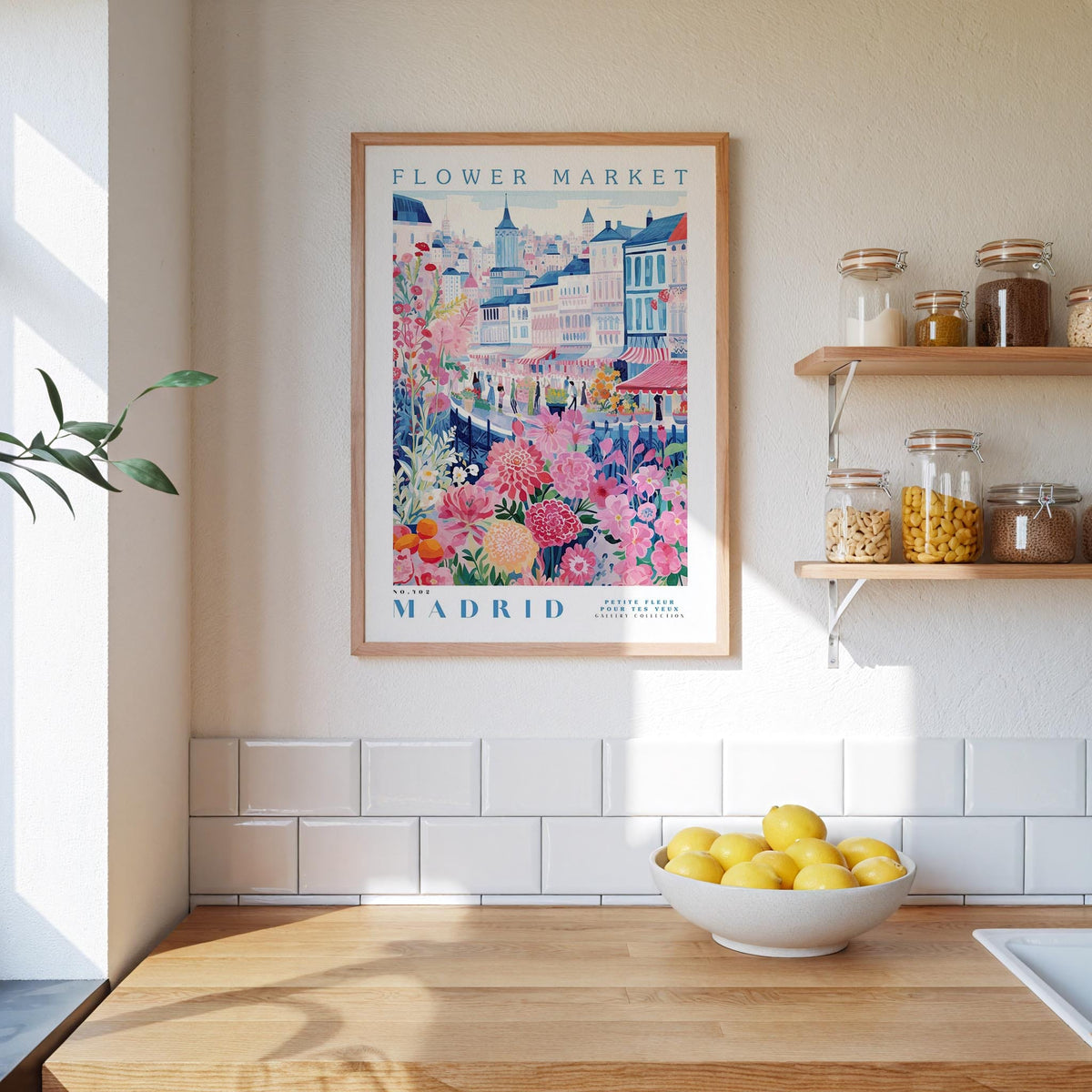 a framed poster of a colorful illustration of a flower market in Madrid, Spain, hanging on a wall above a wooden counter with a bowl of lemons.