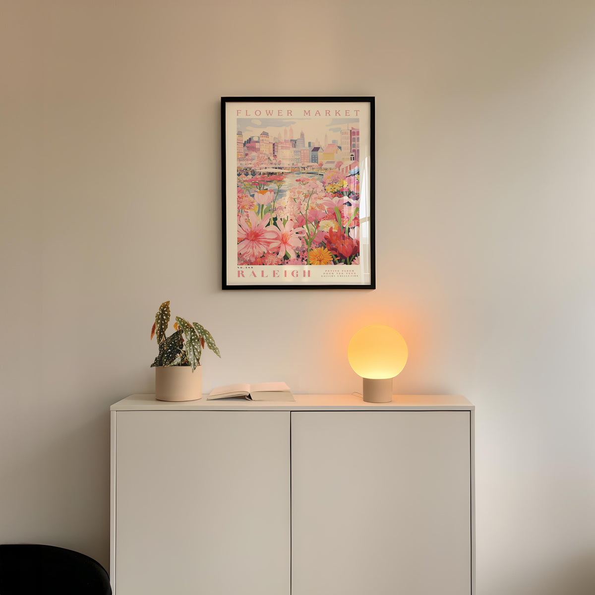 a white cabinet with a potted plant and a lamp on top, set against a wall with a framed poster of a cityscape above it.