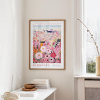 a framed poster of a colorful floral scene hanging on a wall, with a white table and a vase of flowers nearby.