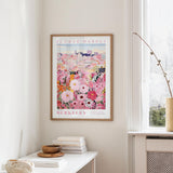 a framed poster of a colorful floral scene hanging on a wall, with a white table and a vase of flowers nearby.