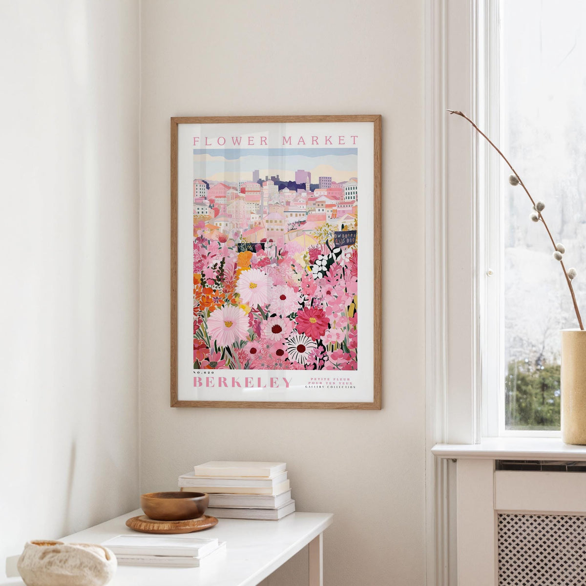 a framed poster of a colorful floral scene hanging on a wall, with a white table and a vase of flowers nearby.