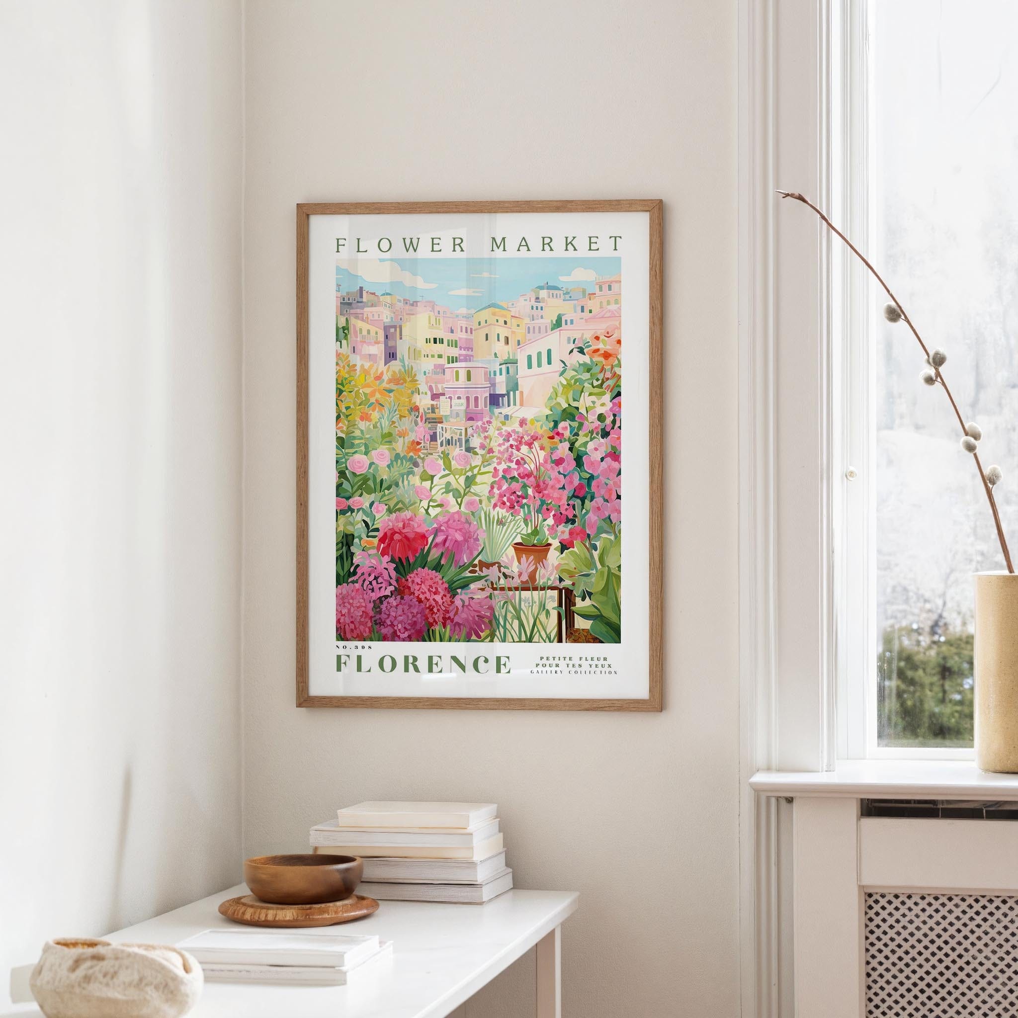 a framed poster of a colorful garden scene hanging on a wall, with a white table and a vase of flowers in the foreground.