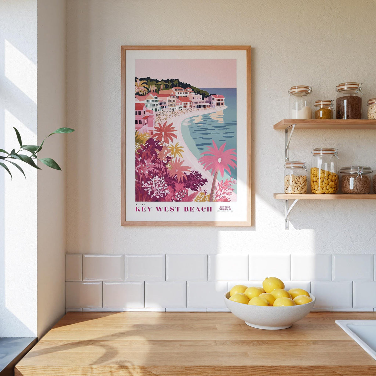 a framed poster of a tropical beach scene with a small town, hanging on a wall above a kitchen counter with a bowl of lemons and jars of food.
